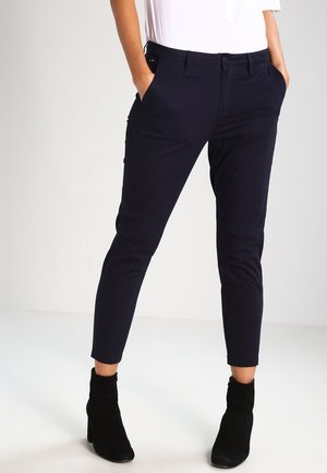Person trägt eng anliegende marineblaue, knöchellange Hose, weißes Oberteil und schwarze, mit Absatz versehene Stiefeletten, steht mit Händen in den Taschen.