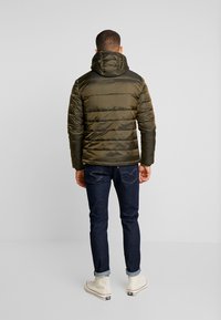 Zelená olivová zateplená bunda s kapucí, s horizontálními švy a hladkou texturou, nošená přes tmavě modré džíny a bílé tenisky.