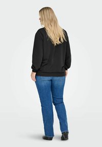 Mujer con cabello rubio largo, vestida con un suéter negro, jeans azules y zapatos negros, de pie de espaldas contra un fondo liso.