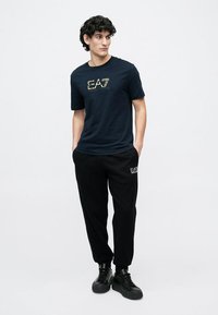 Junger Mann mit lockigem Haar, der ein marineblaues EA7-T-Shirt, schwarze EA7-Sweatpants und schwarze Sneakers trägt, steht mit den Händen in den Taschen.