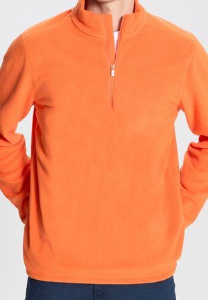 Man draagt een feloranje fleece trui met een kwart ritssluiting en lange mouwen, staand met de handen achter de rug.