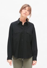 Black long-sleeve shirt featuring a button-up front, two chest pockets, and a relaxed fit. Smooth texture and collar design are prominent.