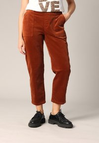 Pantalon en velours côtelé de couleur rouille chaude, avec deux poches avant, coupe droite et texture douce. Associé à des chaussures noires.