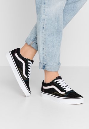 Osoba ubrana w jasnoniebieskie podwinięte jeansy oraz czarne niskie trampki z białymi sznurowadłami i wielokolorowym przeszyciem, stojąca na białej podłodze.