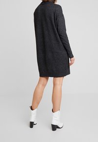 Vestito a maglia grigio scuro fino al ginocchio con maniche lunghe, texture morbida, spacchi laterali e polsini a costine, indossato con stivali con tacco bianchi alla caviglia.