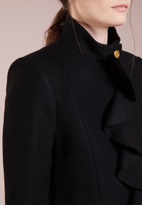 Cappotto di lana nero con colletto alto, caratterizzato da un evidente dettaglio arricciato e un accento con bottone dorato al collo. Texture liscia, design aderente.