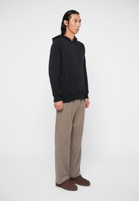 Felpa nera con cappuccio e tasca frontale, abbinata a pantaloni larghi marroni texturizzati e scarpe marroni scure. Design semplice e colori neutri.