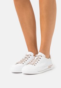 Baskets blanches avec une tige en cuir texturé, lacets beige et un détail au talon en tan contrastant. Semelle en caoutchouc avec de subtils rayures horizontales.