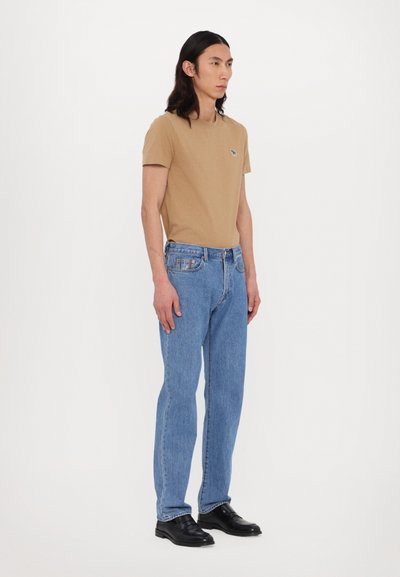 Tan cotton t-shirt with a small embroidered logo, paired with blue denim straight-leg jeans and black leather shoes, standing pose.