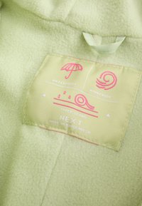 Étiquette de vêtement sur tissu vert pâle montrant les icônes imperméable, coupe-vent et coutures collées avec le texte « NEXT Made in Bangladesh ».