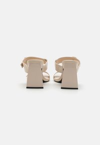 Beige High-Heelsandalen mit breiten Riemen und offenen Zehen, ausgestattet mit einem quadratischen Absatz und strukturiertem Material. Flache schwarze Sohlen runden das Design ab.