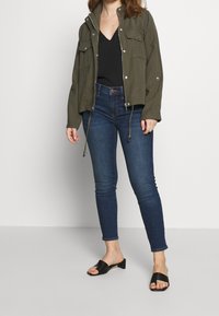 Olivgrön kort jacket med silverdetaljer, två bröstfickor, svart topp, mörkblå figursydda jeans och svarta klacksandaler.