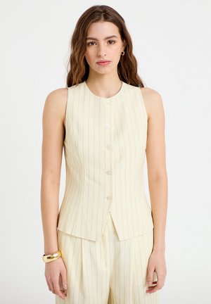 Nainen, jolla on aaltoilevat ruskeat hiukset, pukeutunut hihattomaan kermaiseen raitaneuletakkiin, jossa on napit edessä, ja siihen sopiviin laskostettuihin housuihin. Asusteina kultainen rannekoru ja renkaanmuotoiset korvakorut.