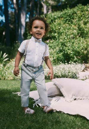 Toddler boy wearing light outfit with suspenders and bow tie stands on grass near outdoor cushions and greenery in daylight.