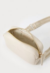Sac en cuir beige à bandoulière avec zip et quincaillerie dorée. L'intérieur est doté d'une doublure en tissu. Sangle ajustable avec détail de logo.