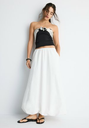 Mladá žena v čierno-bielom topu bez ramienok, dlhej bielej sukni a čiernych sandáloch pózuje opretá o bielu stenu so zatvorenými očami.