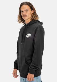 Black hoodie with a front pocket and drawstring hood, featuring a circular white logo on the chest. Made from a soft, textured fabric.