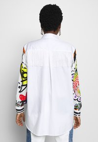 Witte shirt met decoratieve franjes op de achterkant, met kleurrijke graffiti-achtige patronen op de mouwen en gestreepte accenten op de manchetten.