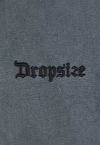 Sivi materijal s izvezenim logotipom "Dropsize" u kontrastnoj crnoj boji. Tekstura je glatka s blagim rastezanjem.