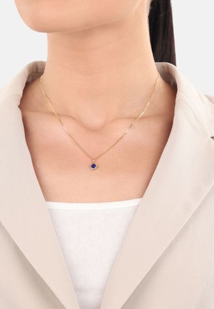 Collana d'oro con un pendente blu rotondo, indossata su un collo nudo sopra un top bianco, completata da un Blazer beige chiaro.
