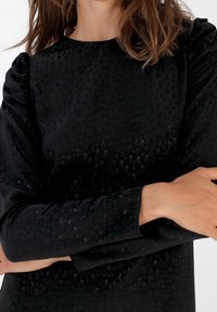 Femme portant un haut noir texturé à manches longues avec des motifs subtils d'étoiles et de crânes, bras croisés à la taille sur fond blanc.