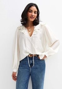 Witte blouse met een gerimpelde halslijn en lange mouwen, gecombineerd met blauwe high-waisted jeans en een bruine riem. Glad materiaal, ontspannen pasvorm.