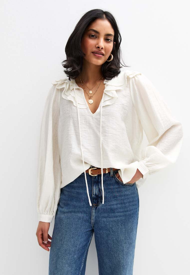 Witte blouse met een gerimpelde halslijn en lange mouwen, gecombineerd met blauwe high-waisted jeans en een bruine riem. Glad materiaal, ontspannen pasvorm.