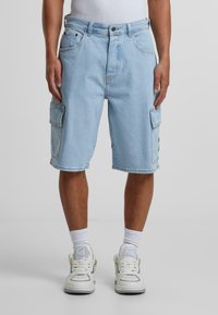 Lyseblå denim cargo-shorts med sidelommer, løs passform og frynset kant, kombinert med hvite joggesko og ankelsokker.