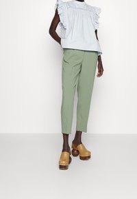 Chemisier bleu clair avec des manches à volants, pantalon vert sur mesure et sabots marron avec des talons en bois et des accents en clous.