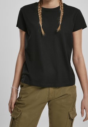 Persona con el pelo rubio trenzado que lleva una camiseta negra lisa de manga corta y pantalones cargo verde oliva con bolsillos.