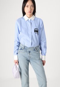 Sinine ja valge triibuline nööpidega särk valge kraega ning musta graafilise printimisega rinna-taskul, seostatuna helesiniste teksade ja lavendlivärvi kotiga.