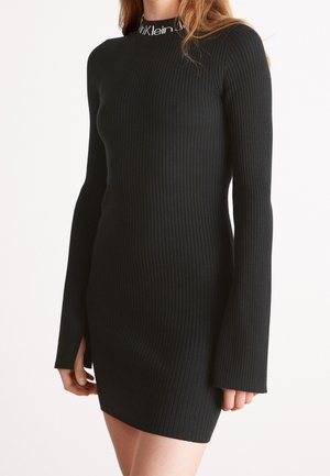 Mujer con un vestido ajustado de manga larga de canalé negro con cuello alto que presenta texto blanco en el cuello.