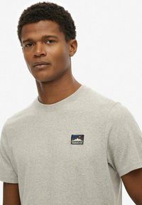 T-shirt in cotone grigio chiaro con un piccolo logo ricamato blu, arancio e bianco sul lato sinistro del petto. Design classico con collo a giro.