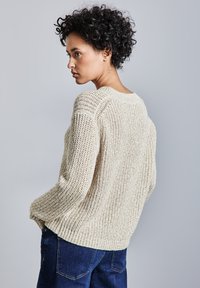 Maglione a maglia textured beige con vestibilità ampia e orlo a coste, abbinato a pantaloni in denim blu scuro. Visibilità del motivo a maglia e dettagli della forma.