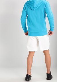 Felpa blu chiaro con tasca frontale e cappuccio con cordino, abbinata a pantaloni corti sportivi bianchi e scarpe nere. Visto da dietro.
