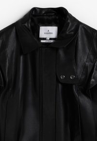 Chaqueta de cuero negro con un cuello tipo solapa, detalles de botones a presión y una textura suave. La etiqueta muestra el nombre de la marca y la información sobre la tela.
