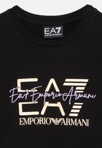 Mustast puuvillast T-särk, millel on metallik-kuldne ja lilla graafiline logo, mis kannab teksti „EA7 Emporio Armani” erinevates stiilides. Ümar kaelus.