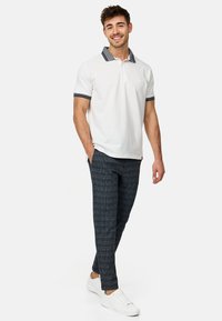 Hvid polo shirt med marineblå stribede krave og mansjetter, parret med marineblå rutede bukser. Model i hvide sneakers, står afslappet.