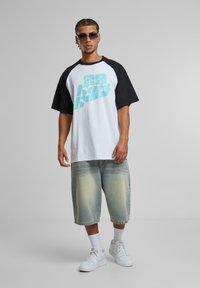 White and black raglan t-shirt with turquoise graphic, paired with light blue denim shorts and white sneakers. Model is wearing sunglasses.