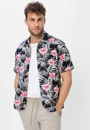 Mann mit kurzen lockigen Haaren, der ein schwarzes Hawaiihemd mit pinken Blumen, ein weißes Unterhemd und hellbeige Hose trägt, die Hände in den Taschen.