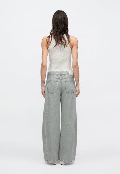 Jean gris à jambe large en denim, avec une taille haute, des poches arrière et une texture lisse. Associé à un débardeur côtelé blanc.
