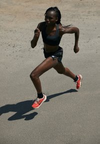 Una corridore in un reggiseno sportivo nero e pantaloni scuri, con capelli intrecciati, che indossa scarpe da ginnastica rosse e bianche, su una superficie grigia texturizzata.