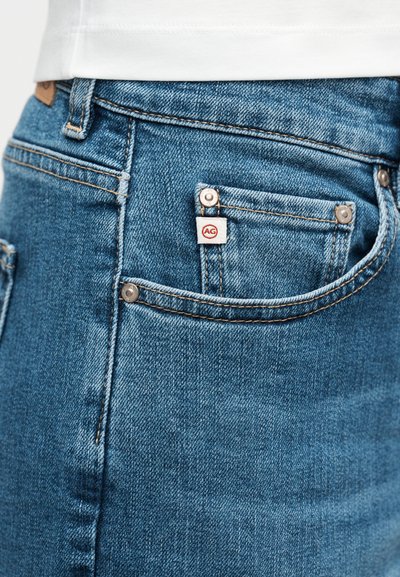 Κοντινή λήψη τσέπης από μπλε τζιν με λευκή και κόκκινη ετικέτα της μάρκας AG, με ορατή ραφή και καρφίτσες, φορεμένη με λευκό πουκάμισο.