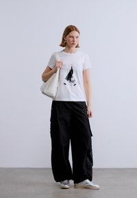 Λευκό μπλουζάκι με εκτύπωση, μαύρο φαρδύ παντελόνι καρό και ασημένια αθλητικά παπούτσια. Το μοντέλο κρατάει μια λευκή τσάντα ώμου σε απλό φόντο.