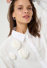 White button-up shirt featuring three textured white fabric roses on the left side, with a classic collar and long sleeves.
