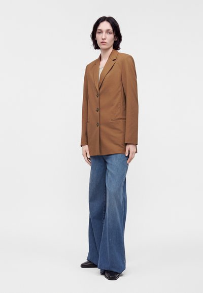 Personne portant un blazer marron boutonné, un jean bleu à jambes larges et des chaussures noires, debout sur un fond clair uni.