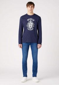 Camiseta de manga larga azul marino con un logo gráfico blanco en el centro, combinada con jeans de mezclilla azules y zapatillas blancas. Diseño sencillo y moderno.
