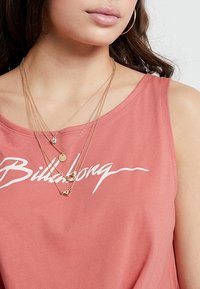 Rosé tanktop med "Billabong"-logo, suppleret med lagdelte guldkæder med cirkulære og trekantede vedhæng. Glat tekstur.