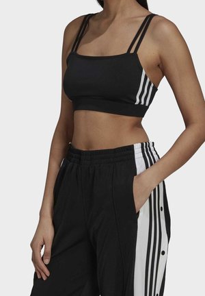 Kobieta ubrana w czarny sportowy biustonosz i czarne spodnie dresowe z białymi paskami po bokach i zatrzaskami, jedna ręka w kieszeni, na jednolitym tle.