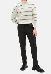 Krémový pruhovaný svetr s vysokým límcem a dlouhými rukávy, kombinovaný s černými tepláky a černými šněrovacími botami.